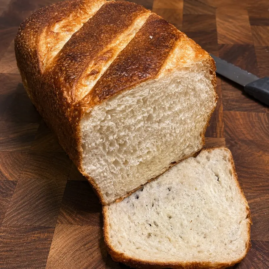 A sourdough sandwich loaf with a scored golden crust, one slice cut to show the soft white crumb, on a wooden board